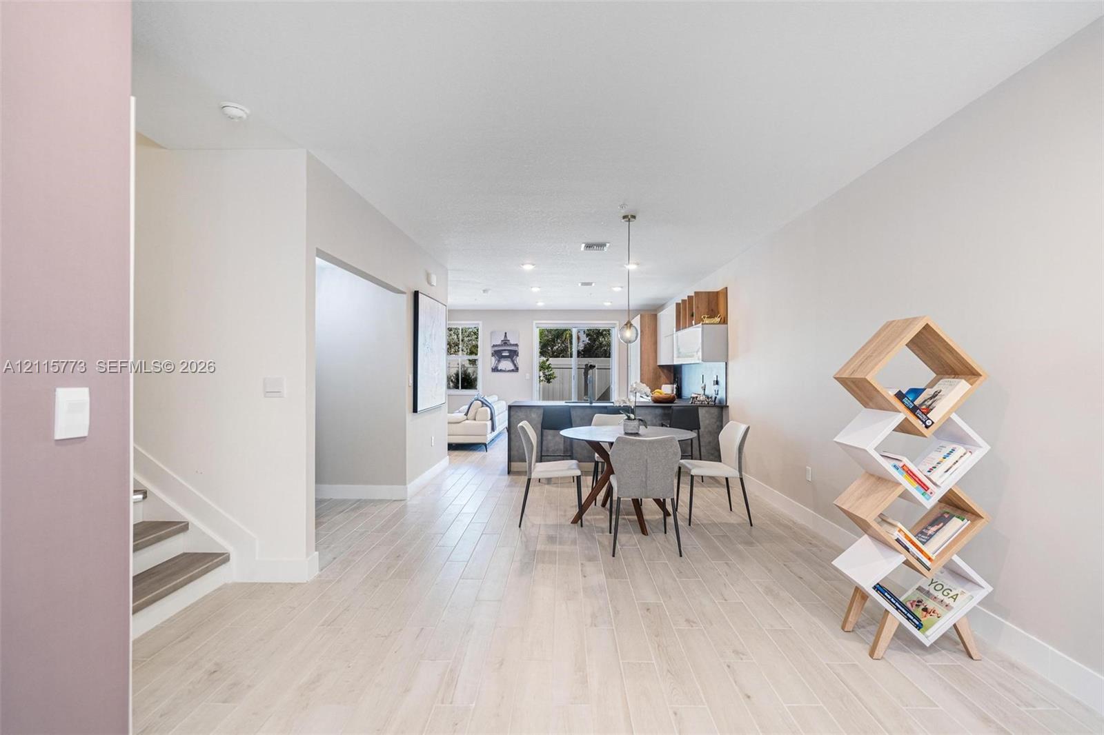 Open-concept interior featuring light wood-finish flooring, recessed lighting, a modern kitchen with dark cabinetry, and a suspended pendant light fixture
