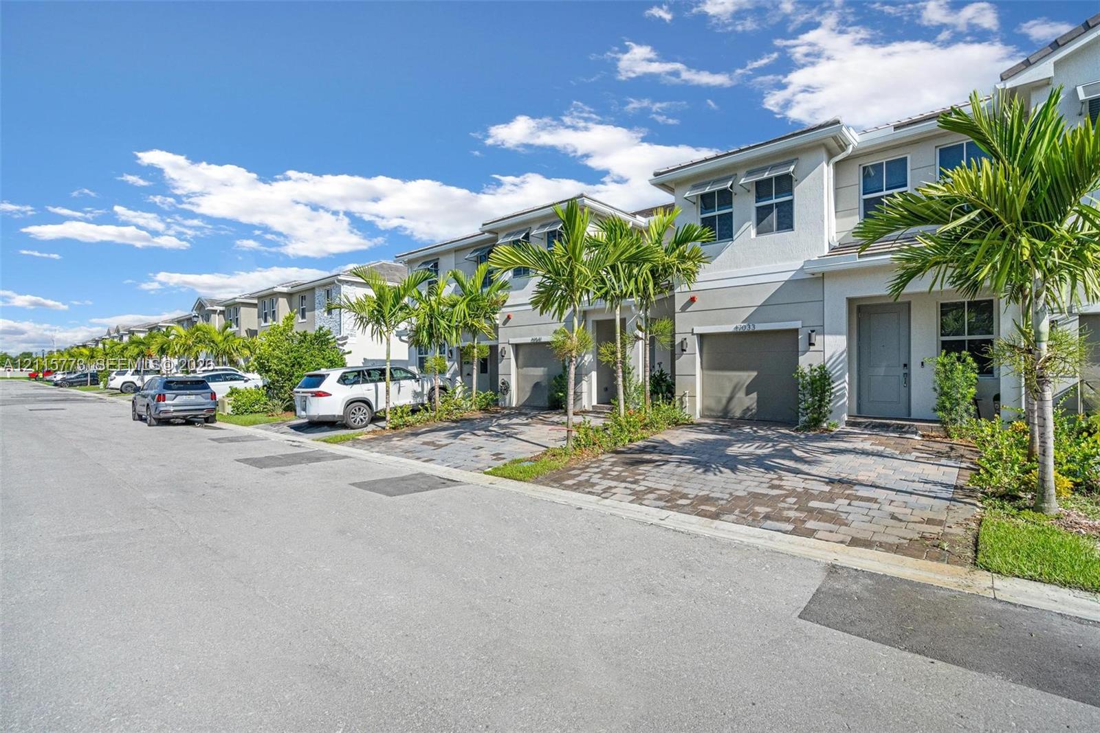 Two-story townhome exterior featuring a stucco facade, paver driveway, single-car garage, and integrated landscaping with mature palm trees Two-story townhome exterior featuring a stucco facade, paver driveway, single-car garage, and integrated landscaping with mature palm trees