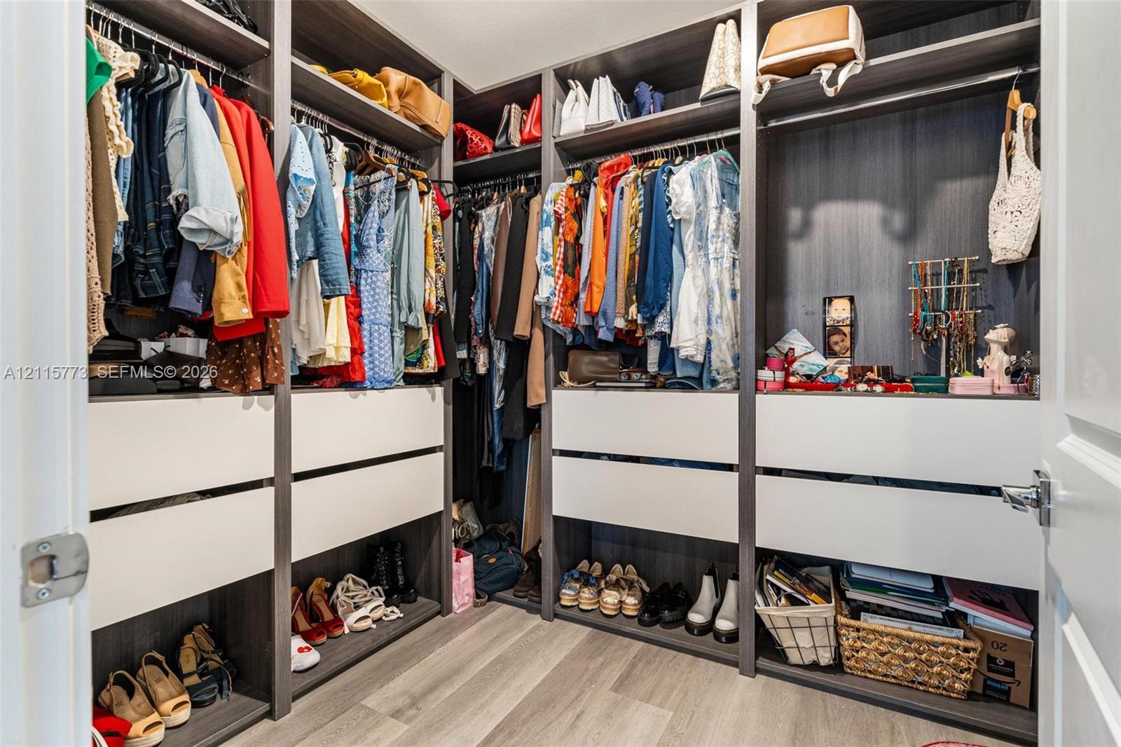 Walk-in closet featuring multi-tiered shelving, built-in drawer units with matte white faces, integrated hanging rods, and wood-finish flooring