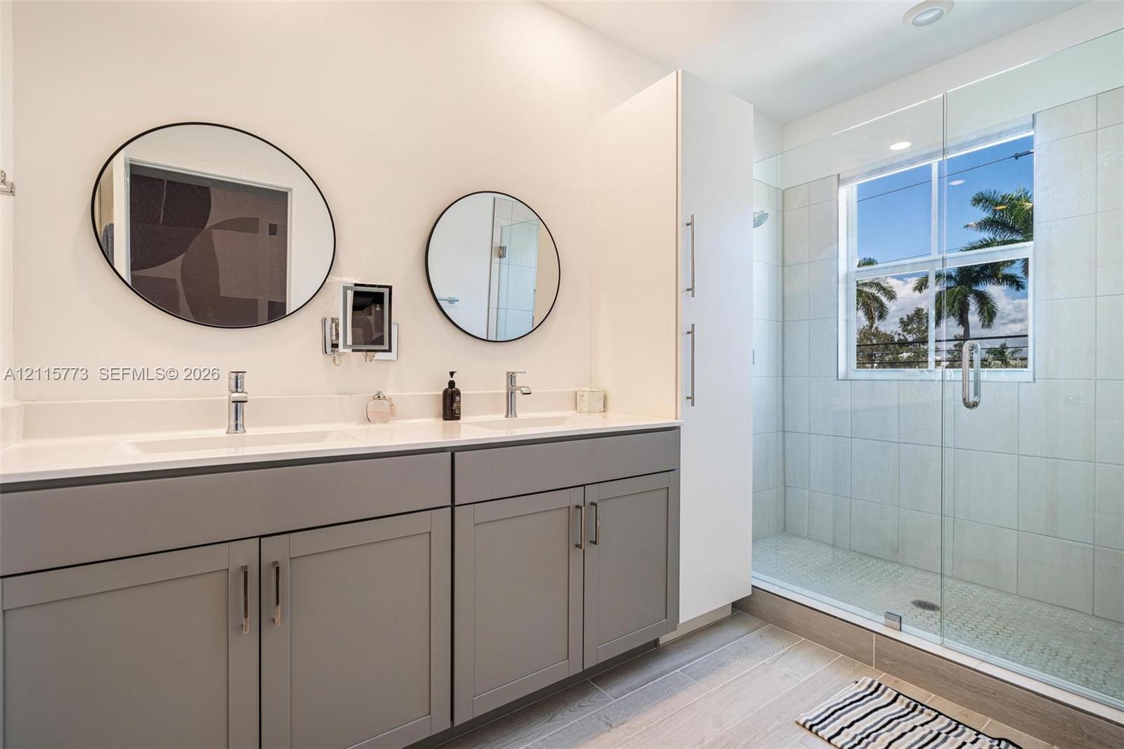 Dual-sink vanity with light-colored countertops, grey cabinetry, wall-mounted circular mirrors, a tall floor-to-ceiling storage cabinet, and a walk-in shower with glass enclosure and light-toned tile surround