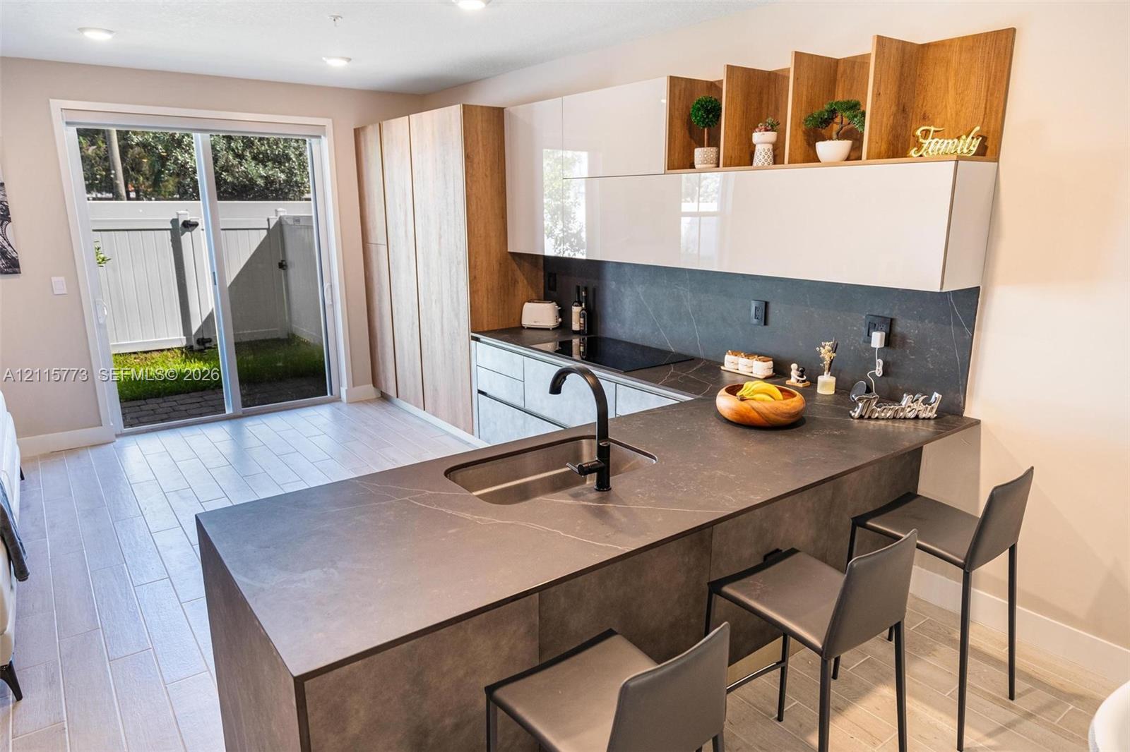 Kitchen featuring two-tone cabinetry with wood-finish accents, matte-finish countertops with integrated sink, dark faucet, built-in cooktop, and glass sliding doors leading to a patio area
