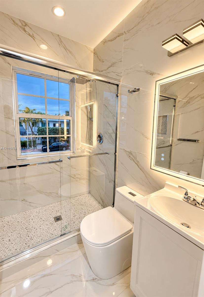 Full-height stone-look wall tile, walk-in shower with pebble-finish flooring and frameless glass enclosure, backlit LED vanity mirror, white cabinetry with integrated sink, and recessed lighting