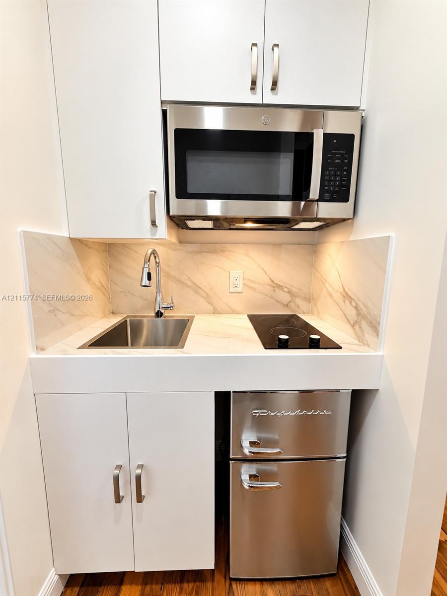 Compact kitchenette featuring white cabinetry, stone-finish backsplash and countertop, stainless steel sink with pull-down faucet, glass cooktop, and over-the-range microwave