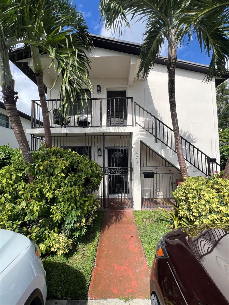 Two-story exterior featuring a white stucco facade, black metal stairwell railings, a second-level balcony, and mature palm trees