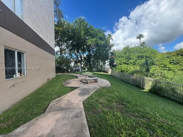 Ground-level exterior area featuring a concrete walkway, a stone-finish circular table with benches, manicured lawn, and perimeter metal fencing
