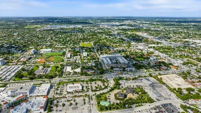 Aerial perspective featuring expansive urban infrastructure, multi-level commercial structures, paved parking facilities, and surrounding green space