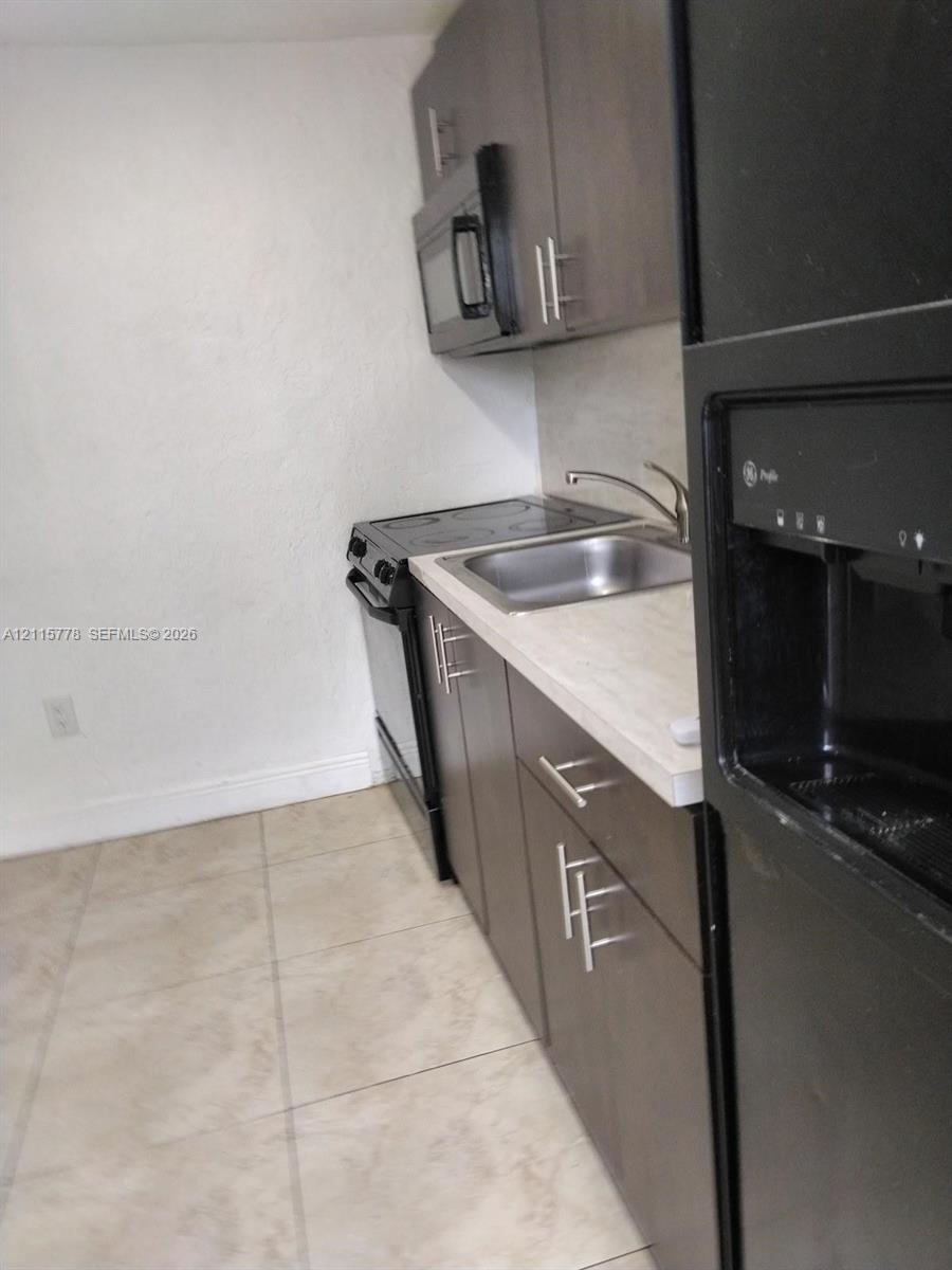 Kitchenette featuring dark-finish cabinetry with modern hardware, neutral stone-finish countertops, stainless steel sink with gooseneck faucet, built-in microwave, and tile flooring