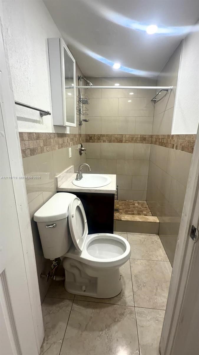 Bathroom featuring a single-sink vanity with a solid-surface countertop, tiled shower enclosure with a decorative accent border, and ceramic tile flooring