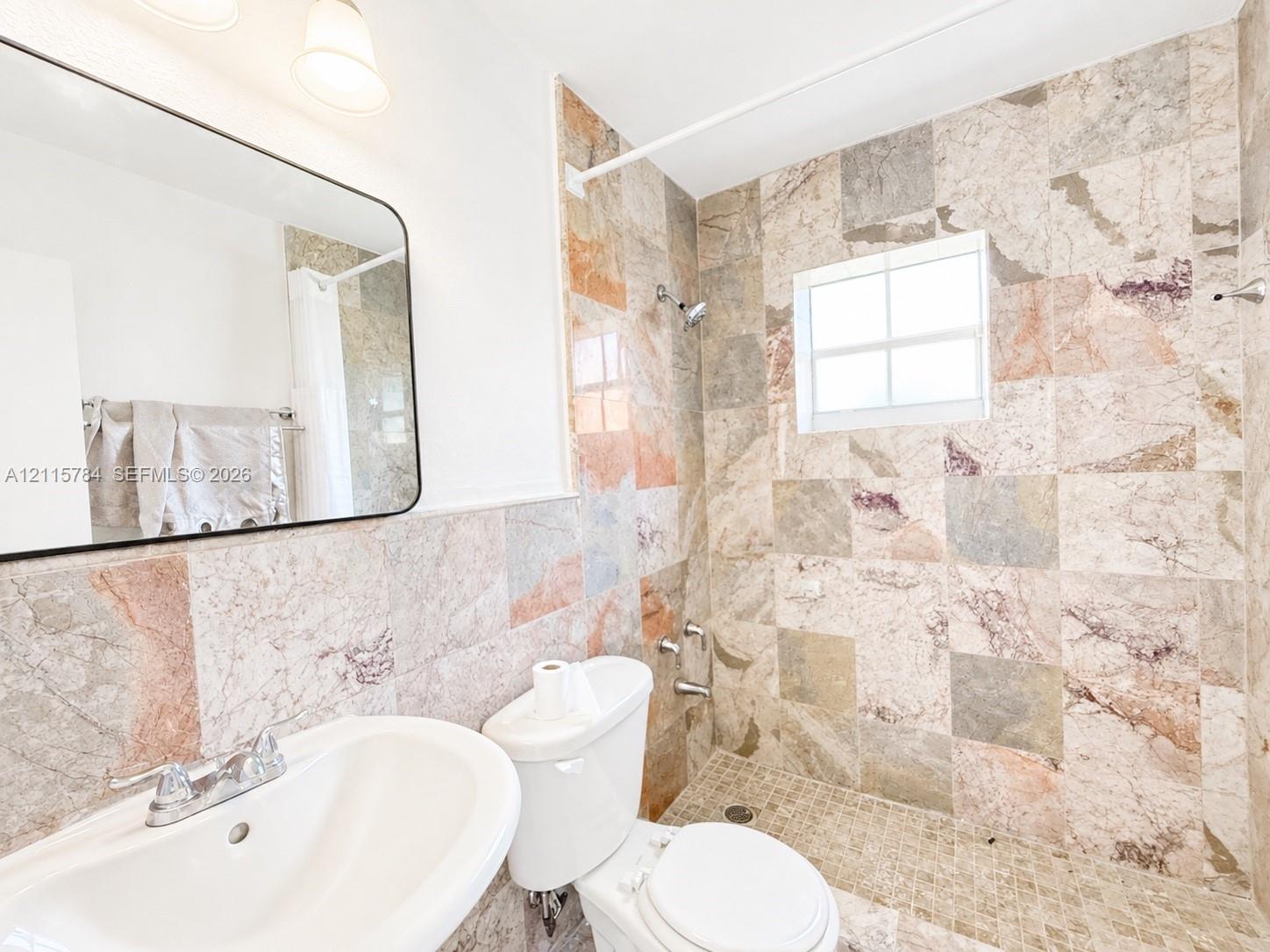Bathroom featuring floor-to-ceiling stone tile, a pedestal sink, a wall-mounted rectangular mirror, and a walk-in shower with stone mosaic flooring