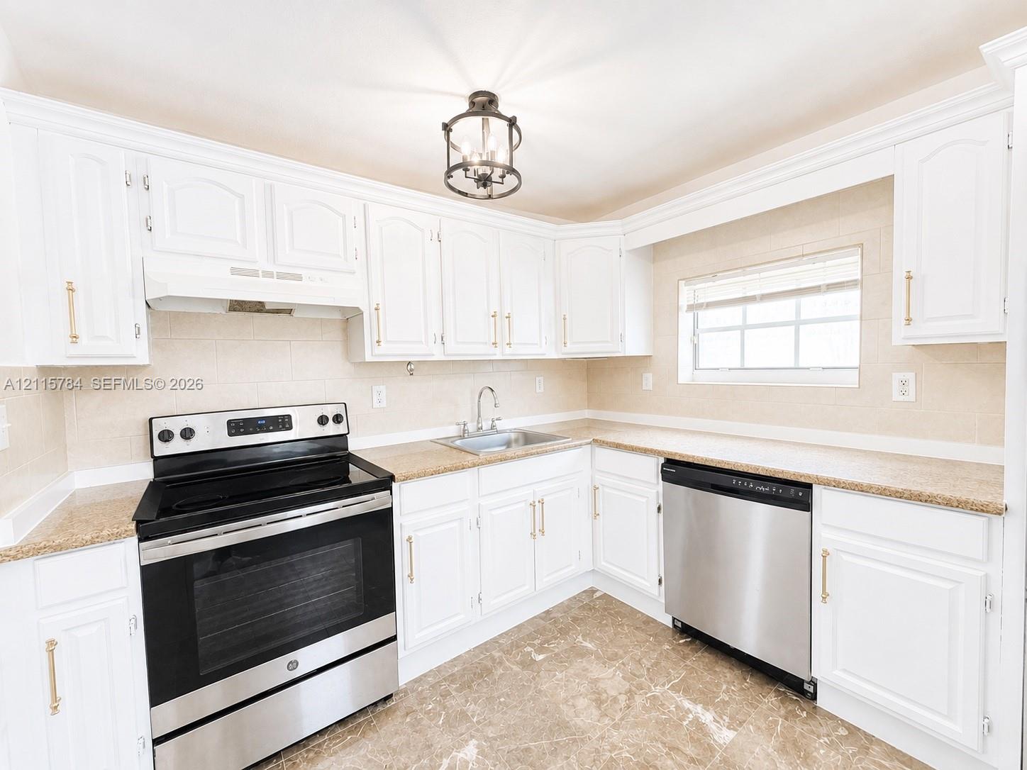 Kitchen featuring white shaker-style cabinetry with gold-tone hardware, stone-finish countertops, stainless steel appliances, stone-tile flooring, and a decorative lantern-style pendant light fixture