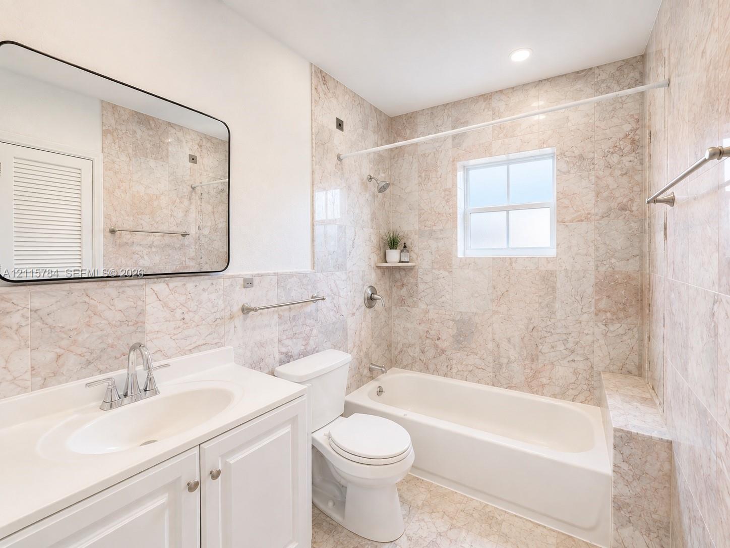 Bathroom featuring full-height stone wall tiling, a built-in bathtub with a shower head, vanity with a white countertop and integrated sink, framed mirror, and a window