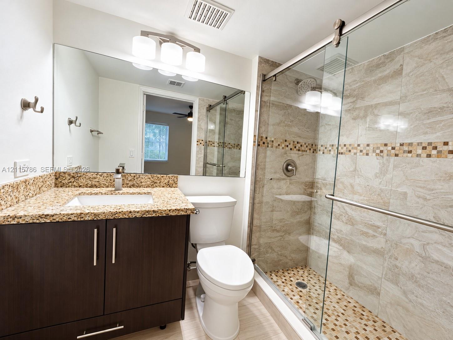 Dark wood-finish vanity with stone-composite countertop, full-width vanity mirror, triple-bulb vanity light, and a walk-in shower featuring glass sliding doors and decorative tile inlay