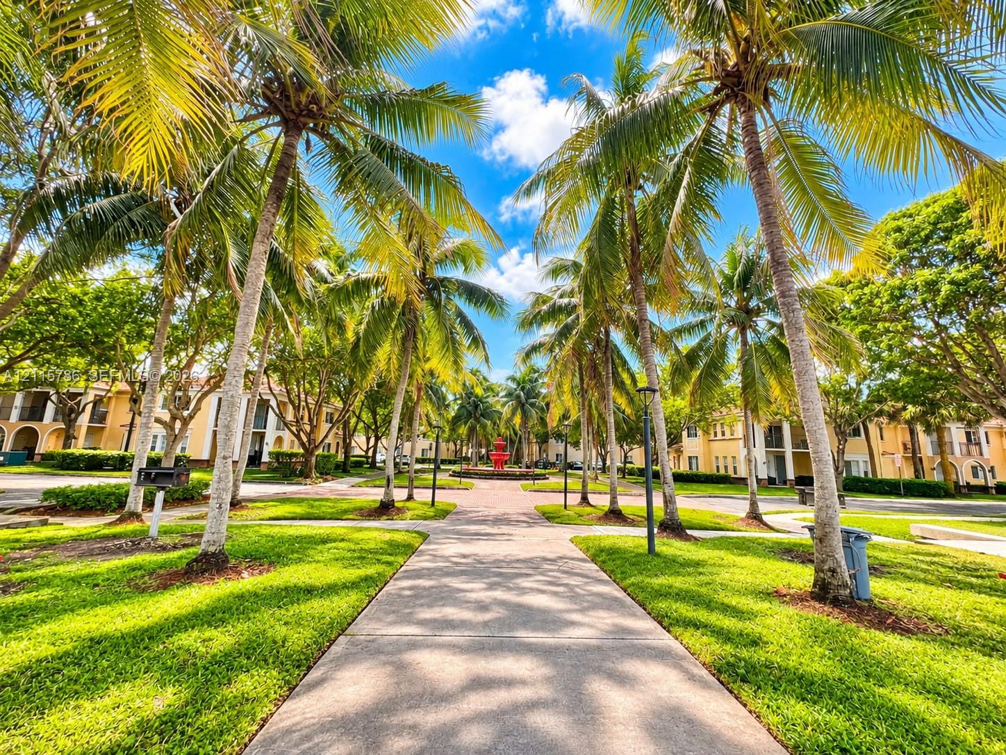 Paved walkway lined with mature palm trees, manicured lawns, and a central fountain