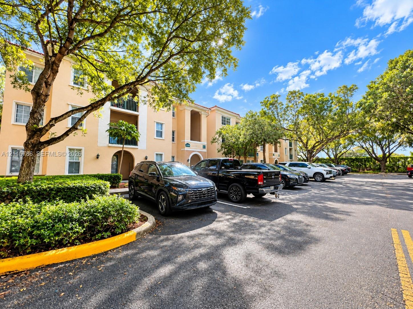 Stucco exterior building featuring balconies, tile roofing, and paved surface parking surrounded by mature landscaping