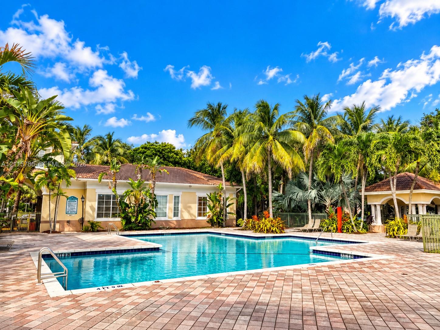 Community swimming pool featuring a brick paver deck, metal pool ladder, built-in pavilion, and mature palm tree landscaping