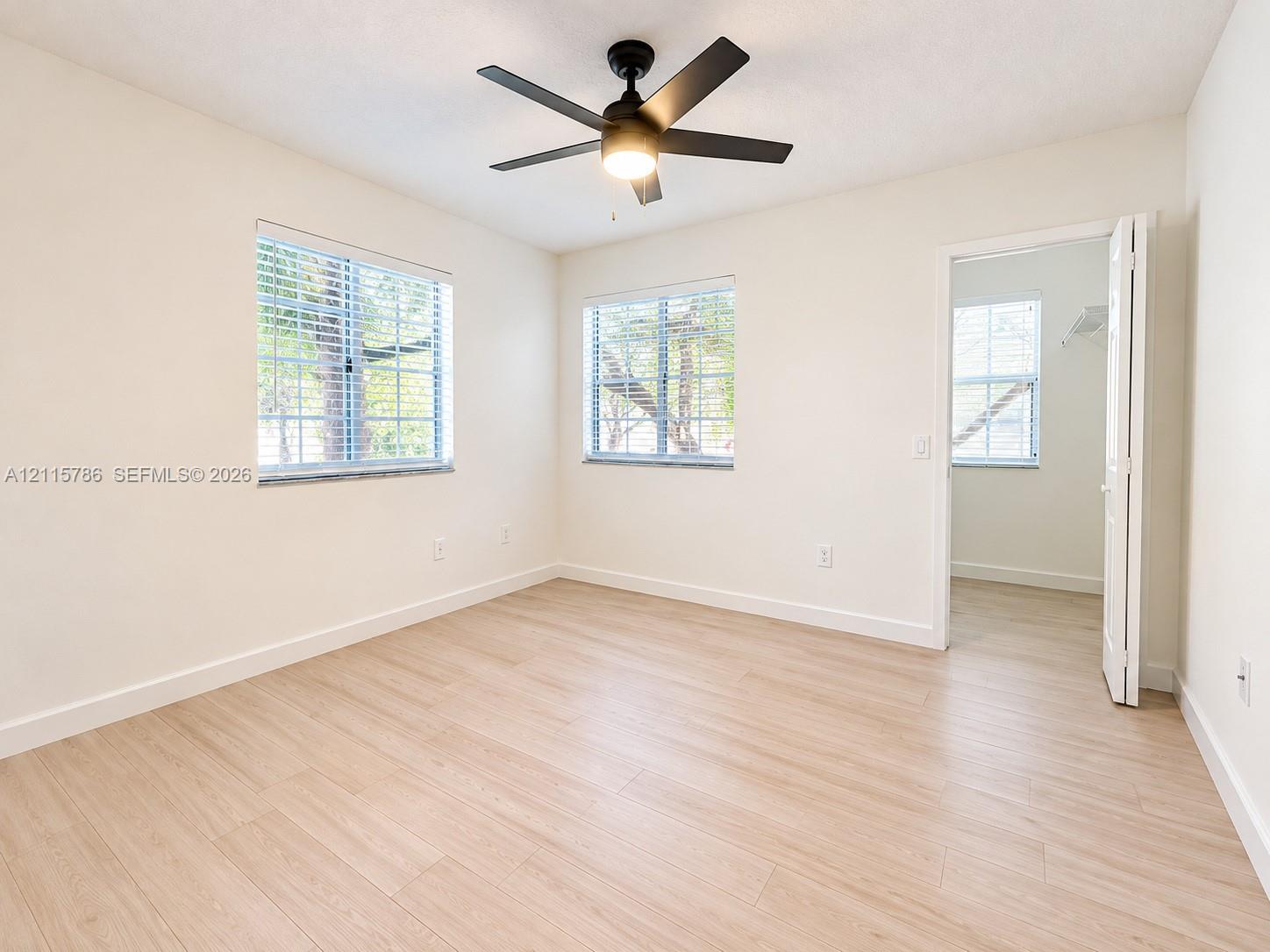 Neutral-toned wood-finish flooring, matte black ceiling fan with integrated lighting, and multiple windows with horizontal blinds