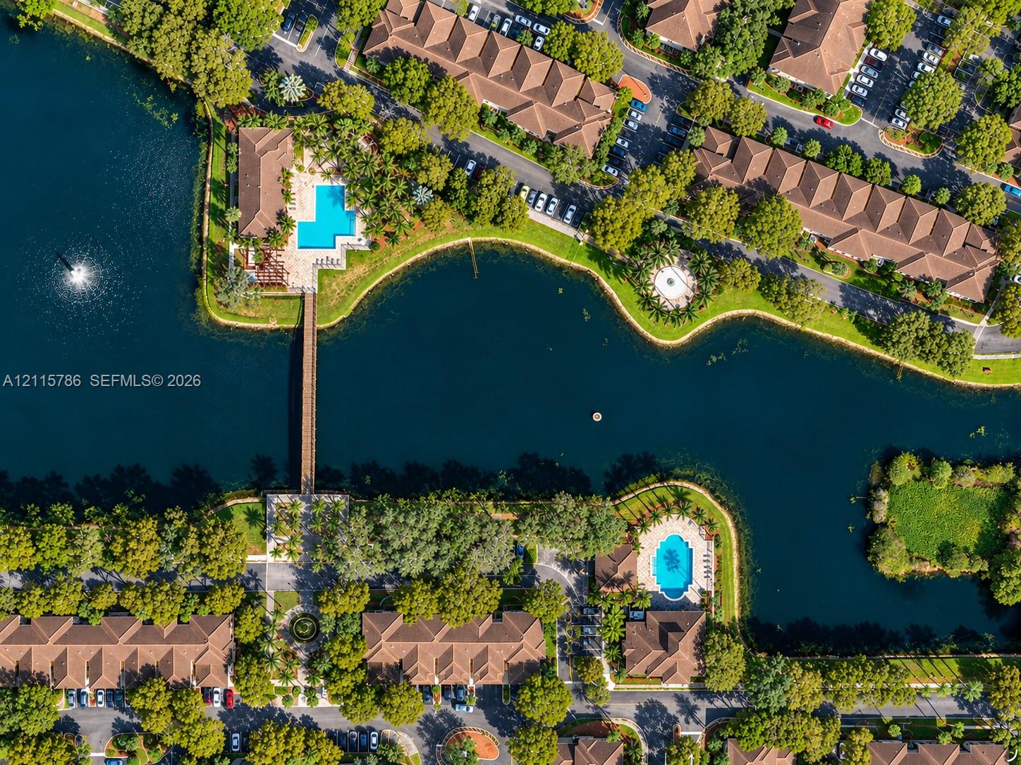 Aerial perspective of a community featuring a central waterway, pedestrian footbridge, swimming pools, and mature trees