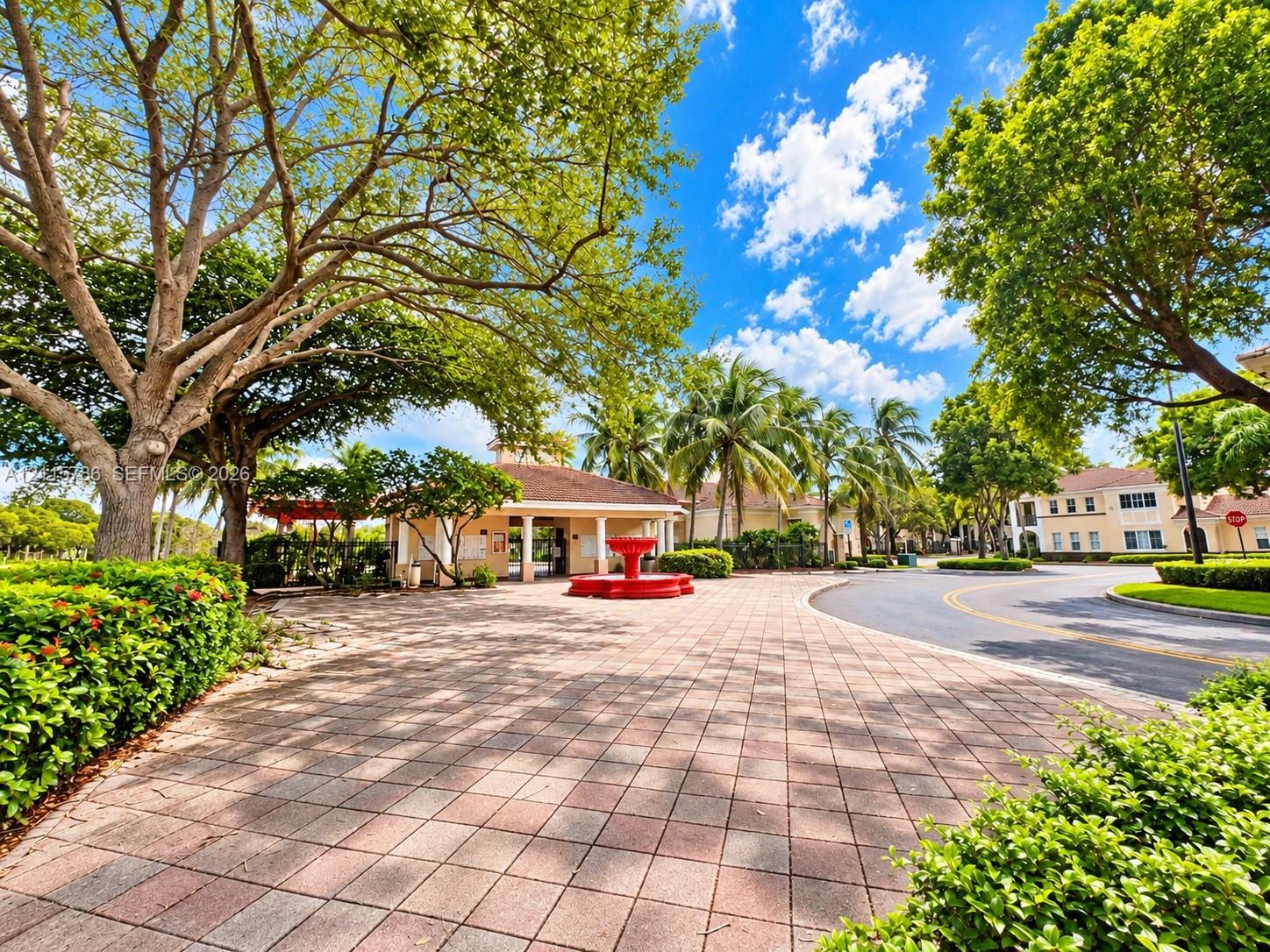 Paver stone entryway featuring a central fountain, mature canopy trees, manicured hedges, and a covered clubhouse structure