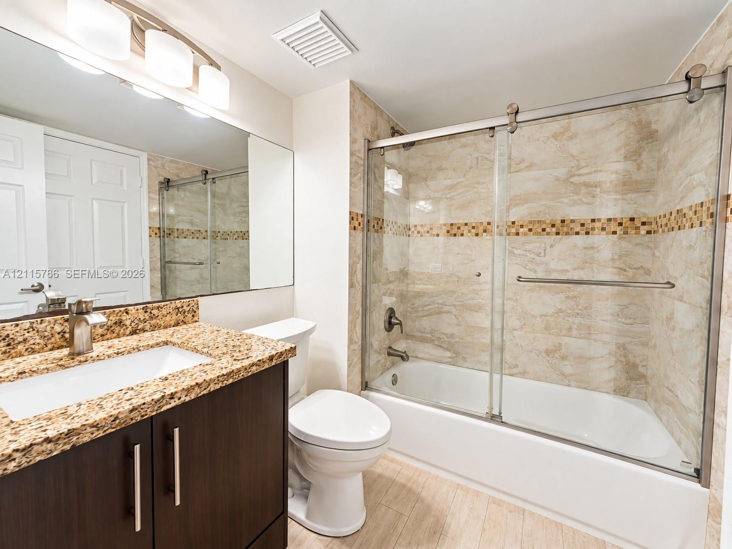 Dark wood-finish cabinetry, stone countertop with an integrated rectangular basin, full-width wall mirror, and a shower-tub combination with sliding glass doors and decorative tile inlay