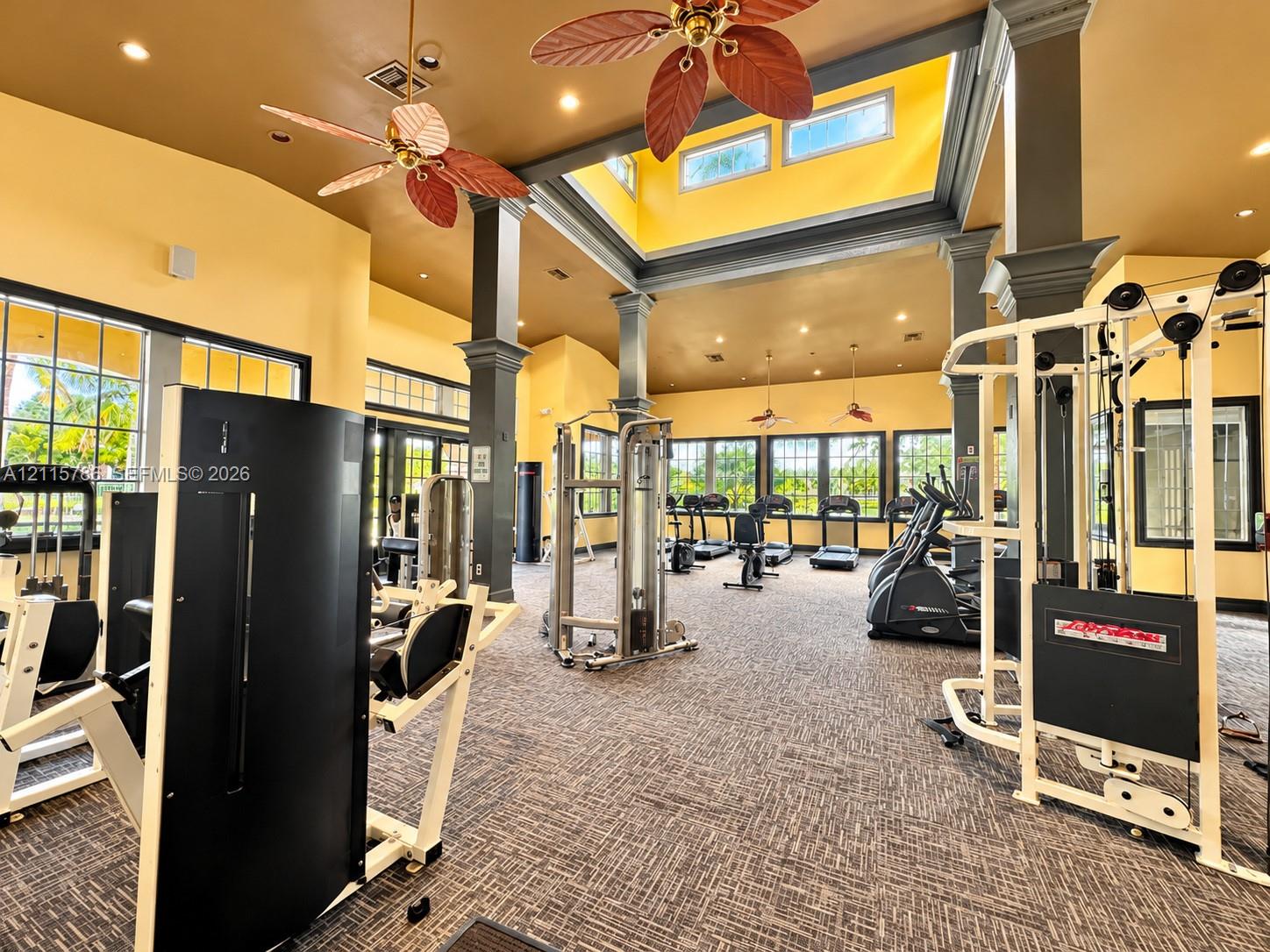 High-ceiling fitness space featuring a central skylight, structural columns with decorative trim, multiple large-pane windows, and inset recessed lighting