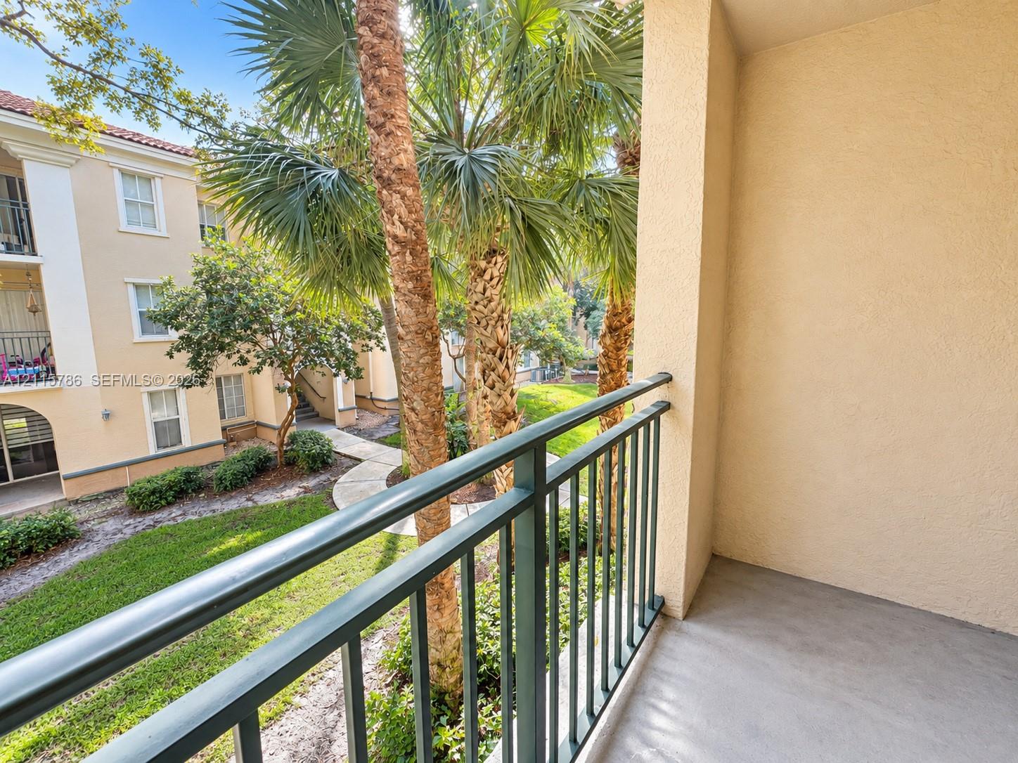 Private balcony featuring textured stucco walls, solid surface flooring, and metal railing overlooking landscaped grounds with mature palm trees