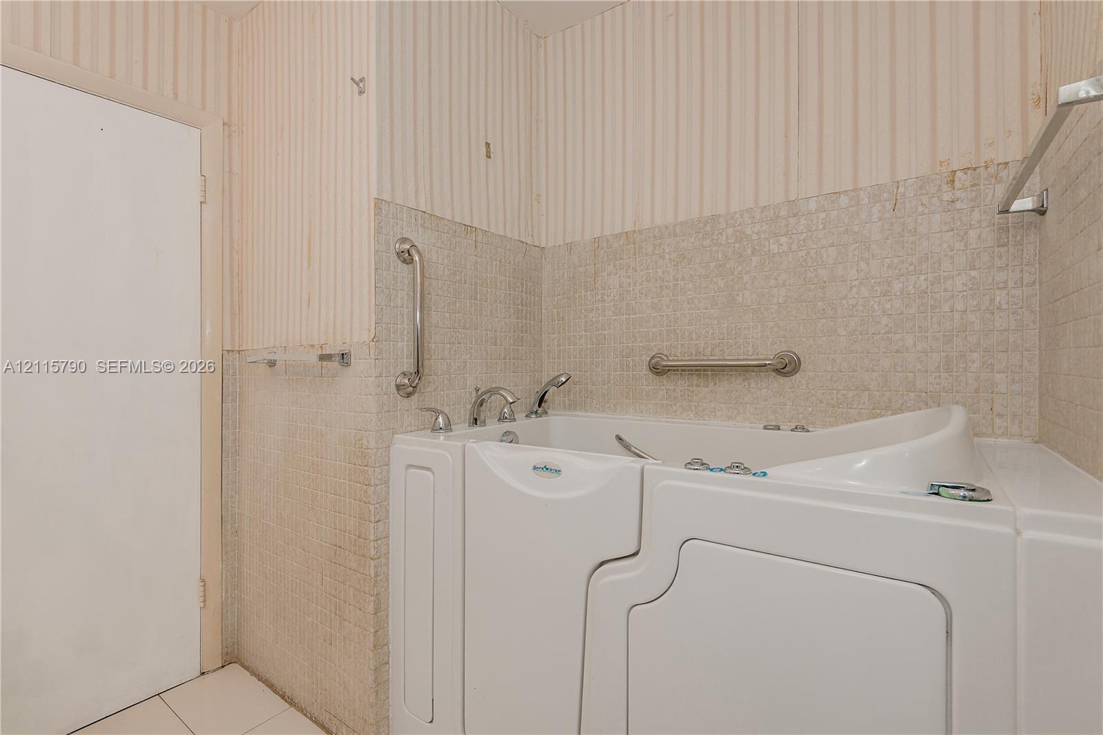Walk-in tub featuring a side-entry door, chrome faucet fixtures, and mounted safety grab bars, complemented by neutral square wall tiling
