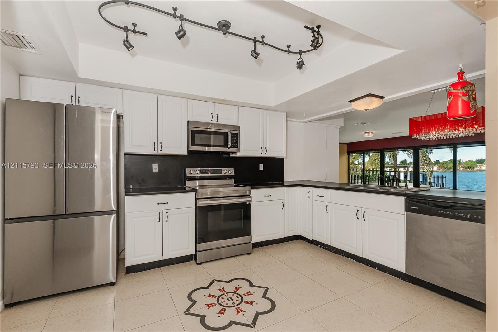 Kitchen featuring white shaker-style cabinetry, black countertops, stainless steel appliances, tile flooring with a central decorative inlay, and adjustable track lighting