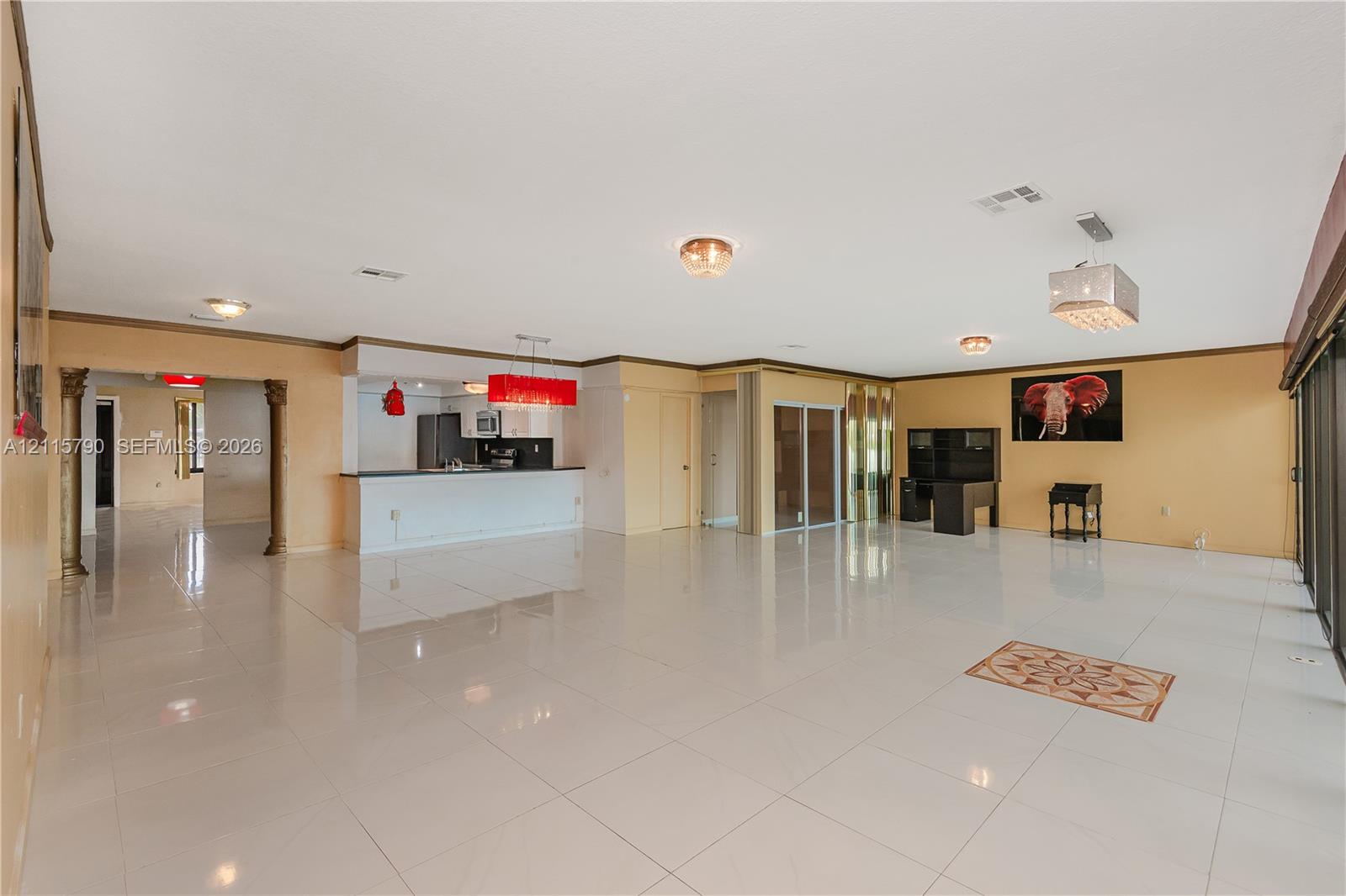 Open-concept interior featuring large-format ceramic tile flooring, built-in breakfast bar with stone-finish countertop, decorative architectural columns, multiple hanging light fixtures, and sliding glass doors