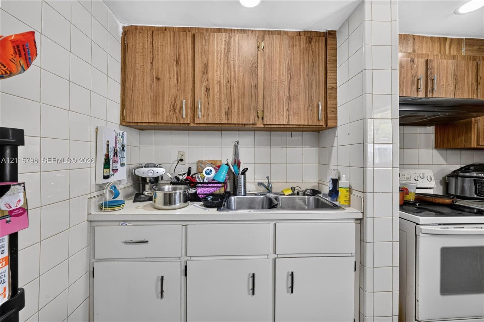 Kitchen area featuring wood-finish upper cabinetry, white lower cabinets, square tile backsplash, and dual-basin stainless steel sink