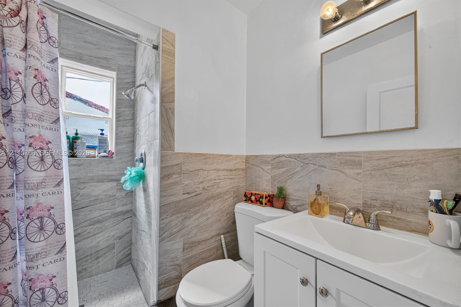 Bathroom featuring a white vanity cabinet with a rectangular basin, chrome faucet, wall-mounted mirror, light fixture, and stone-finish wall tile surrounding the walk-in shower