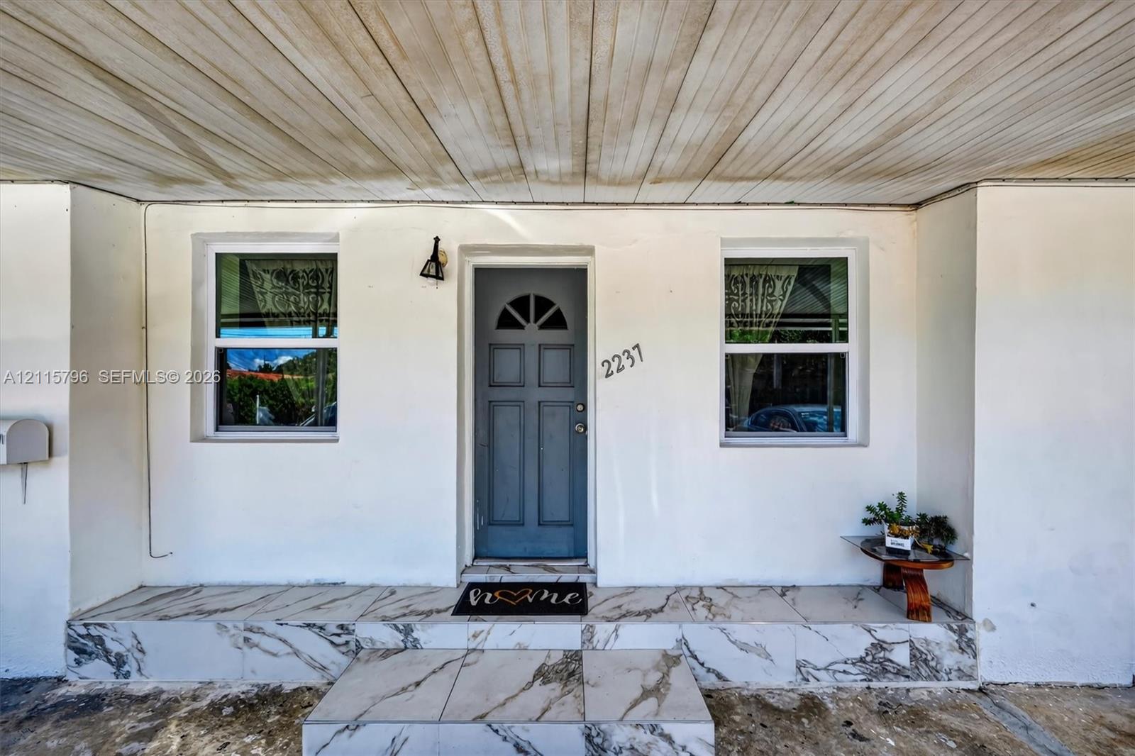 Covered entryway featuring a solid-panel door with a semi-circle transom window, white stucco facade, large-format tile flooring with a marbled finish, and a wall-mounted exterior light fixture
