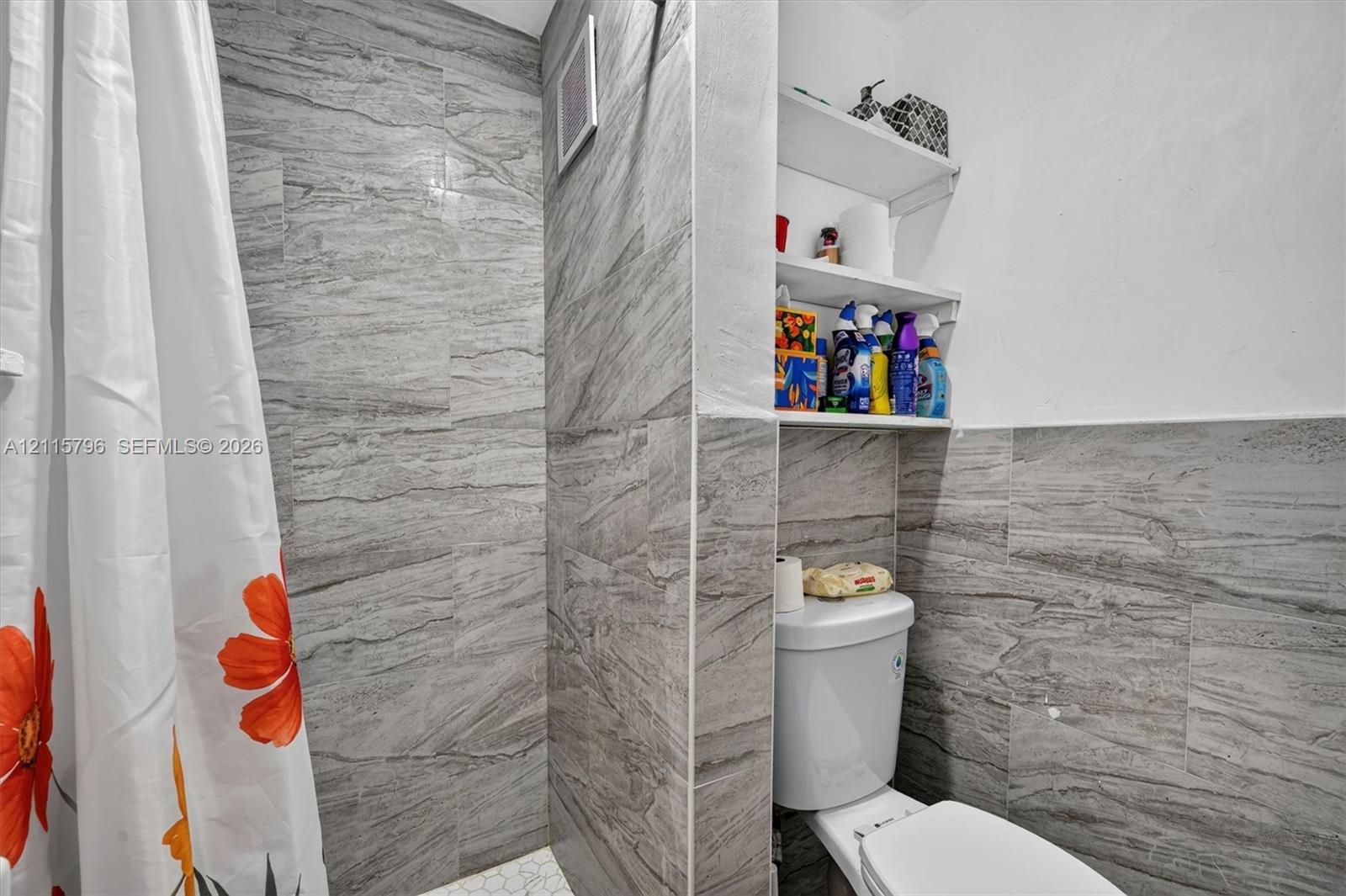 Full-height wall tiling in a grey stone-finish, shower surround, built-in shelving, and a two-piece toilet