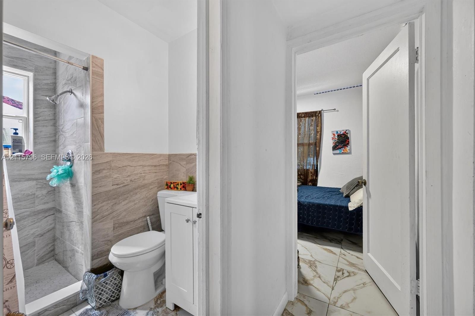 Bathroom featuring a walk-in shower with grey stone-style tile surround, beige tile wainscoting, and a white vanity with a porcelain toilet, complemented by large-format marble-finish tile flooring