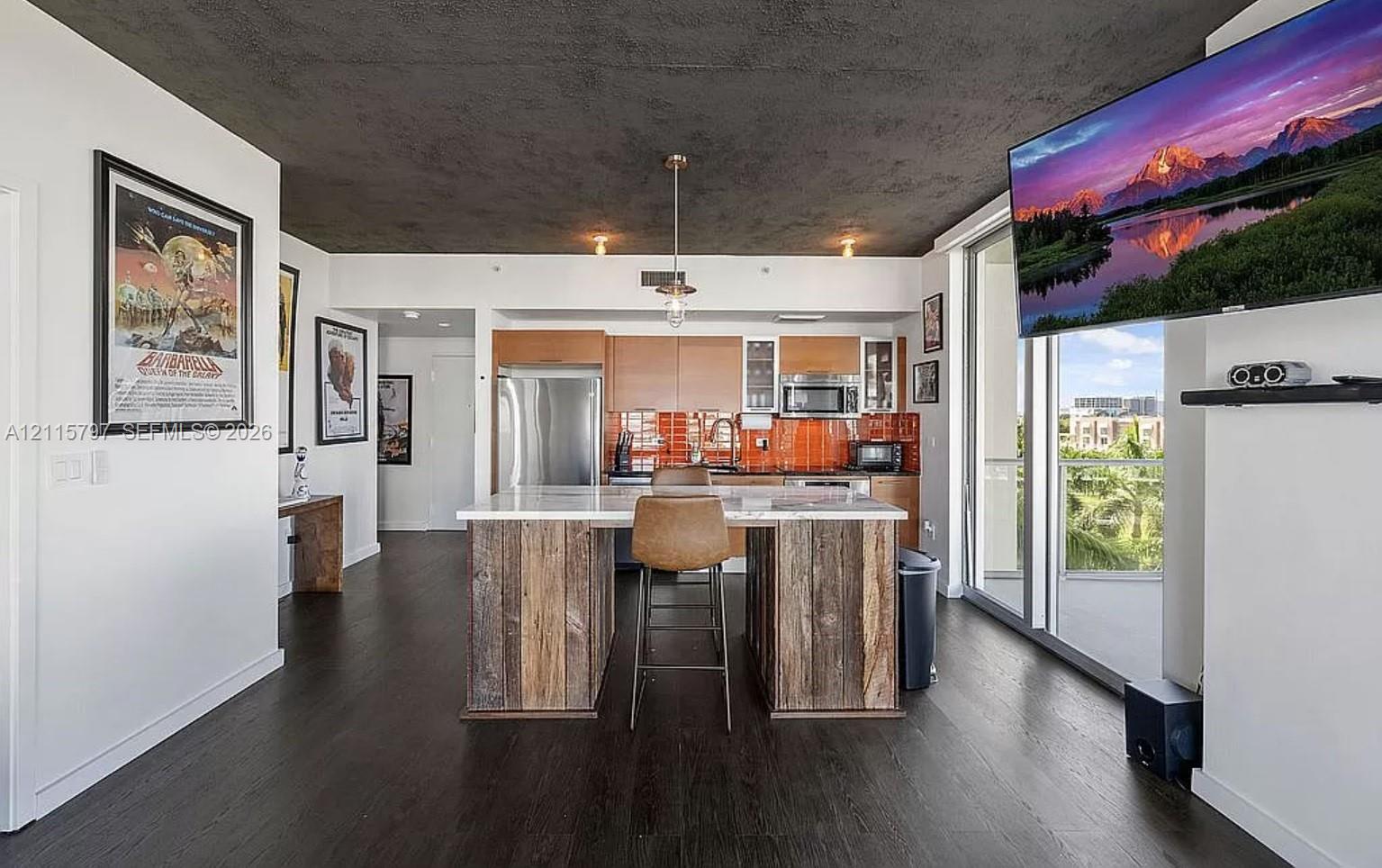 Open kitchen featuring wood-finish flooring, a central island with natural wood cabinetry and a stone countertop, orange glass tile backsplash, and stainless steel appliances Open kitchen featuring wood-finish flooring, a central island with natural wood cabinetry and a stone countertop, orange glass tile backsplash, and stainless steel appliances