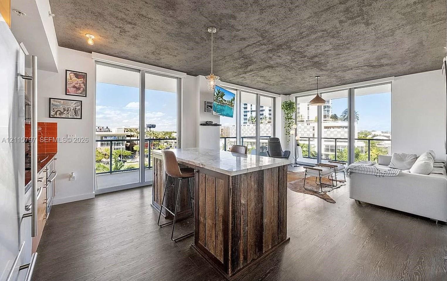 Open-concept living space featuring a textured concrete ceiling, wood-finish flooring, floor-to-ceiling glass doors, and a central island with a stone countertop and reclaimed wood paneling Open-concept living space featuring a textured concrete ceiling, wood-finish flooring, floor-to-ceiling glass doors, and a central island with a stone countertop and reclaimed wood paneling
