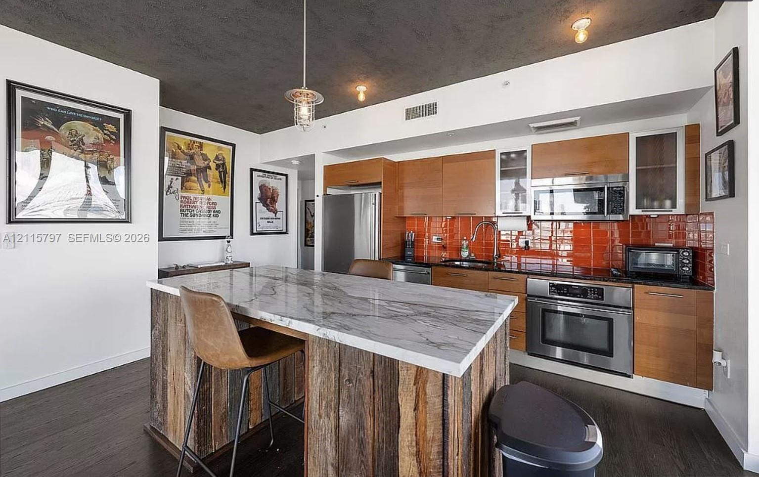 Kitchen featuring flat-panel wood cabinetry, stone-finish countertops, stainless steel appliances, a tile backsplash, and a rustic wood-clad island Kitchen featuring flat-panel wood cabinetry, stone-finish countertops, stainless steel appliances, a tile backsplash, and a rustic wood-clad island