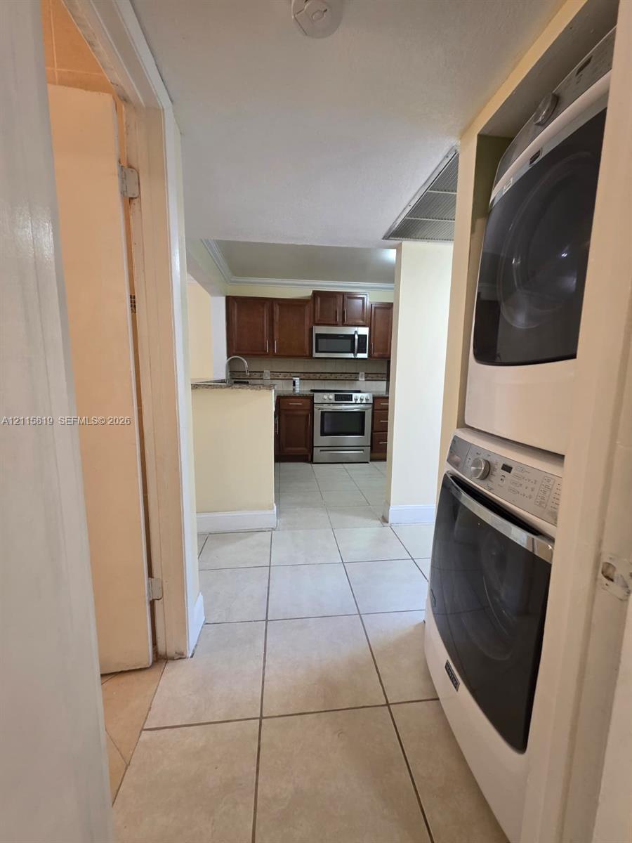Laundry area featuring a stackable washer and dryer, tiled flooring, wood-toned cabinetry, stainless steel appliances, and stone-finish countertops