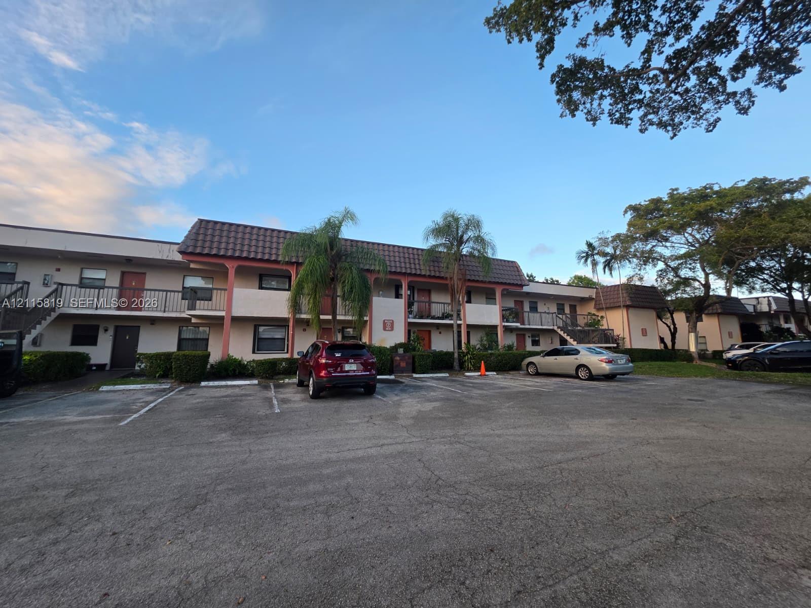 Multi-story exterior featuring a tile roof, paved surface parking, mature palm trees, and ground-level landscaping
