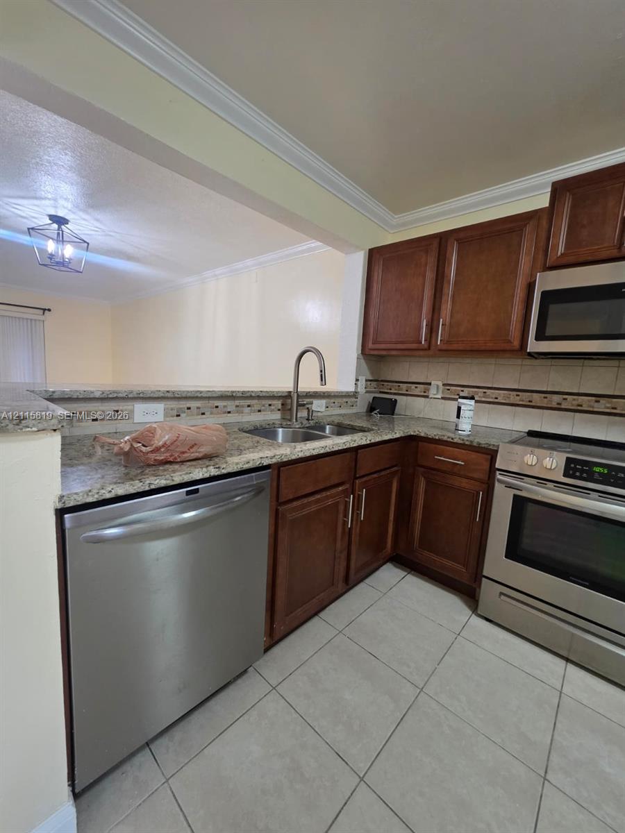 Kitchen featuring medium-toned wood cabinetry, stone-patterned countertops, stainless steel appliances, ceramic tile flooring, and crown molding