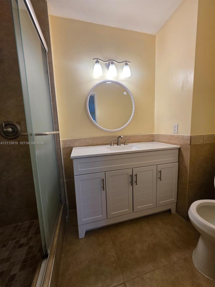 White vanity with rectangular undermount sink, circular framed mirror, three-light wall fixture, and glass-enclosed shower with stone tile flooring