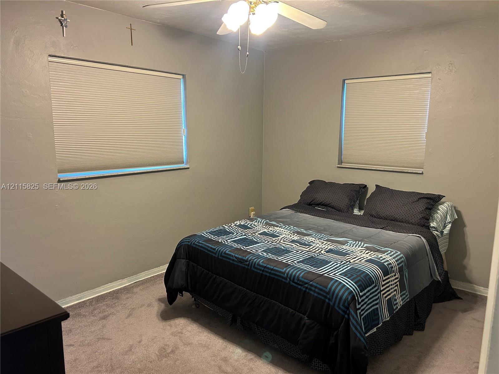 Neutral-toned interior room featuring wall-to-wall carpeting, dual windows with pleated shades, and a central ceiling fan with integrated lighting