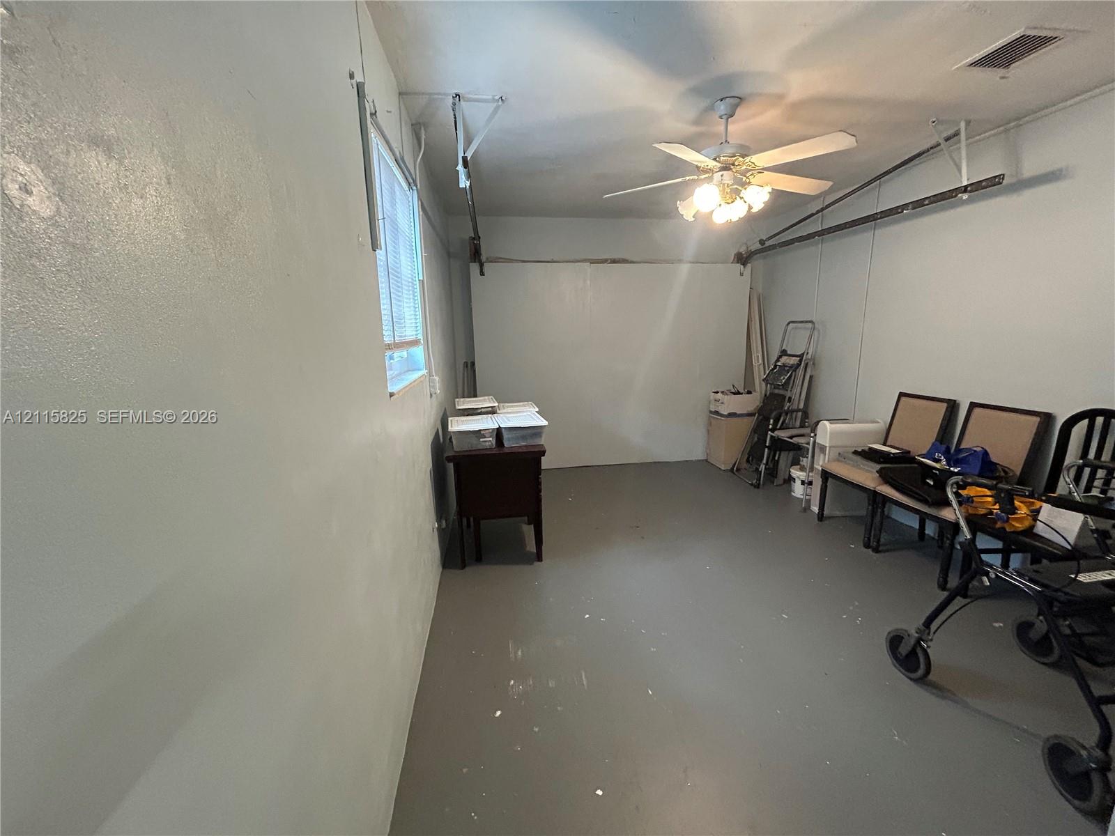 Enclosed garage featuring painted concrete flooring, a single window, and a ceiling-mounted light fixture with a fan