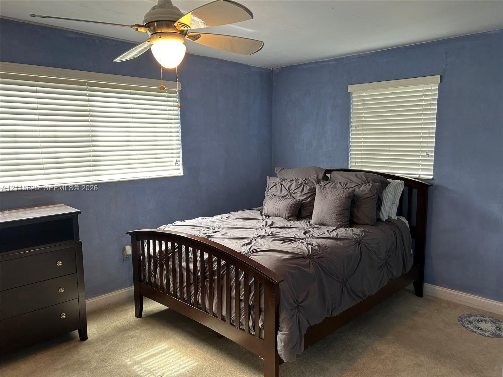 Neutral-toned carpeting, dual windows with horizontal blinds, and a multi-blade ceiling fan with an integrated light fixture
