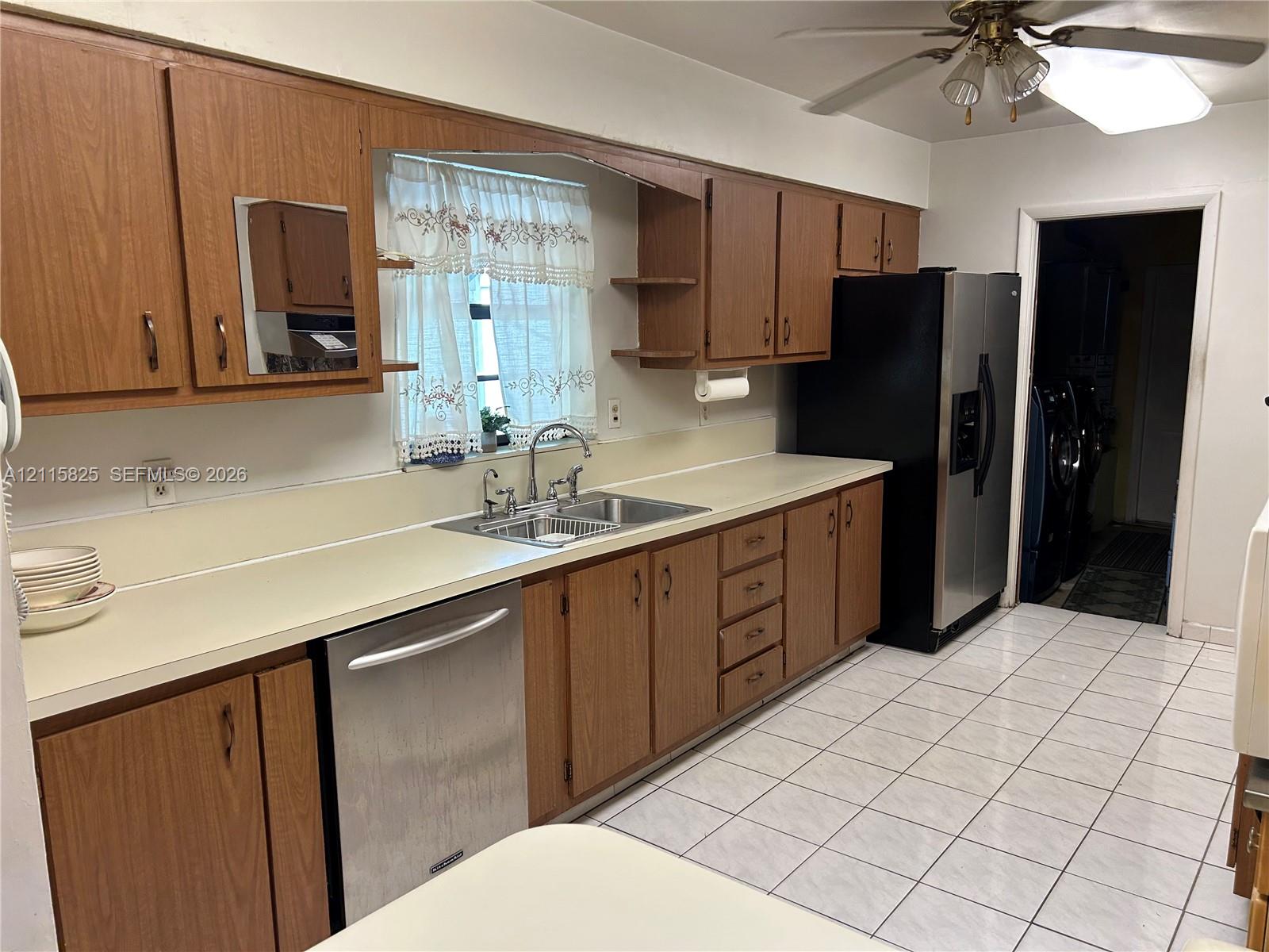 Kitchen featuring wood cabinetry, stainless steel dishwasher, double-basin sink with arched faucet, ceramic tile flooring, and integrated ceiling fan