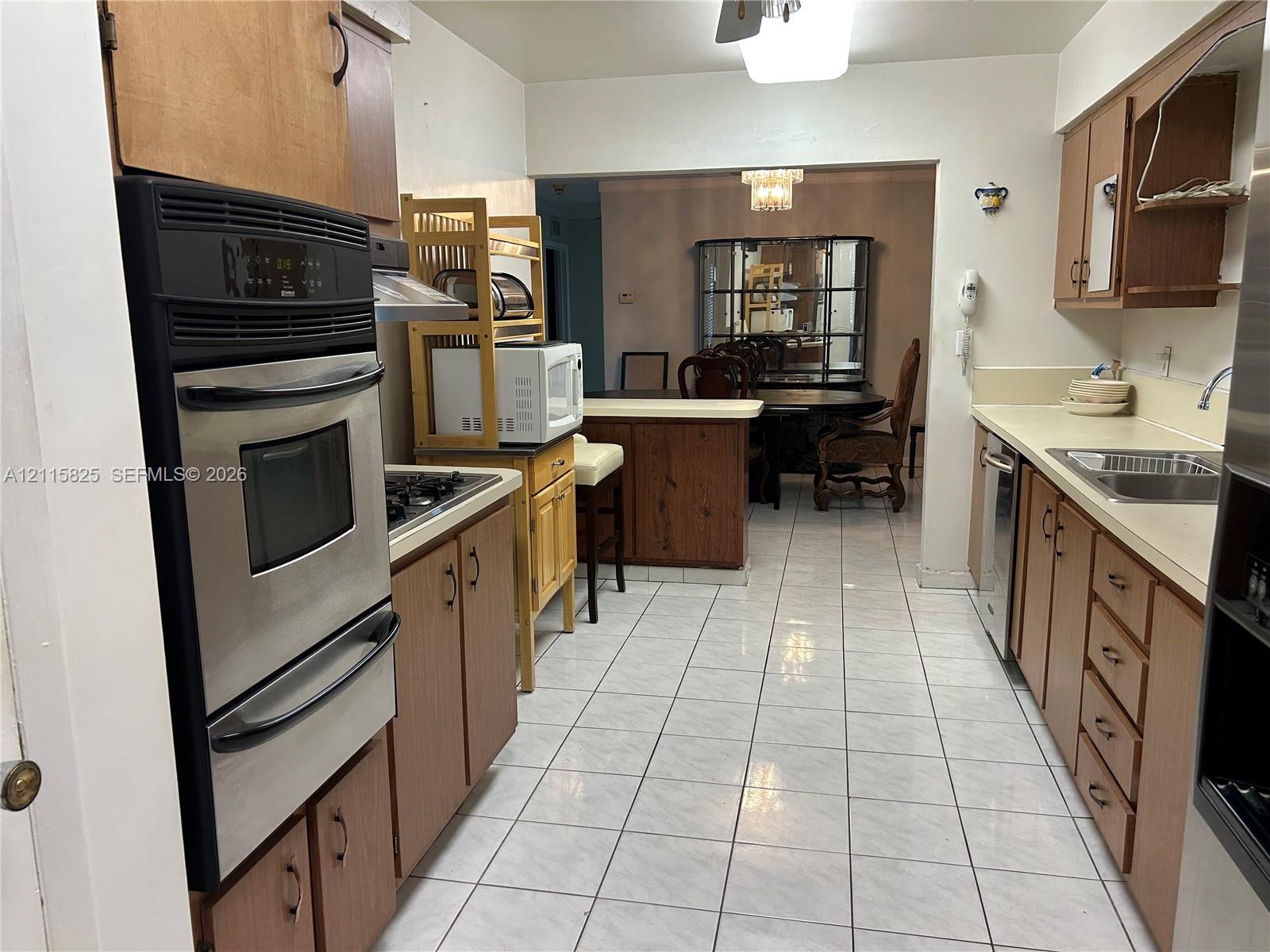 Kitchen featuring wood cabinetry, stainless steel wall-mounted oven, solid surface countertops, a stainless steel dual-basin sink, and tile flooring