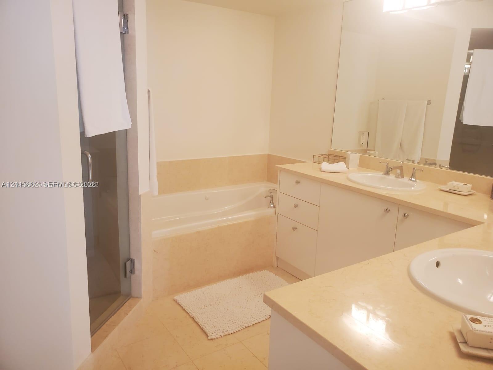 Dual-vanity cabinetry with stone countertops, integrated oval sinks, a large wall-mounted mirror, and a recessed soaking tub with a glass-enclosed shower