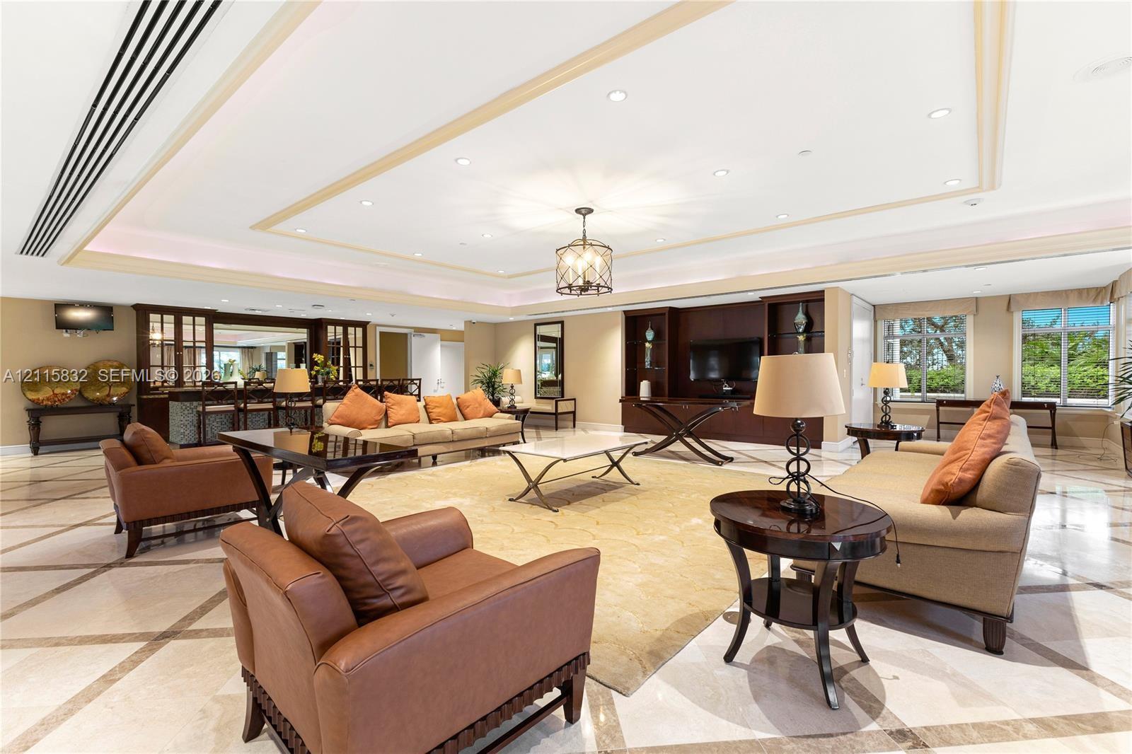 Spacious living area featuring stone tile flooring with a geometric inlay pattern, a tray ceiling with recessed lighting, an integrated wet bar with glass-front cabinetry, and custom built-in shelving