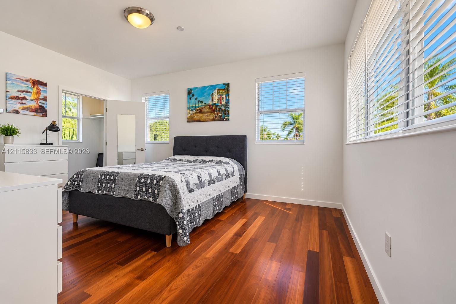 Bedroom featuring wood-finish flooring, white walls, multiple windows with horizontal blinds, and a walk-in closet with a mirrored door