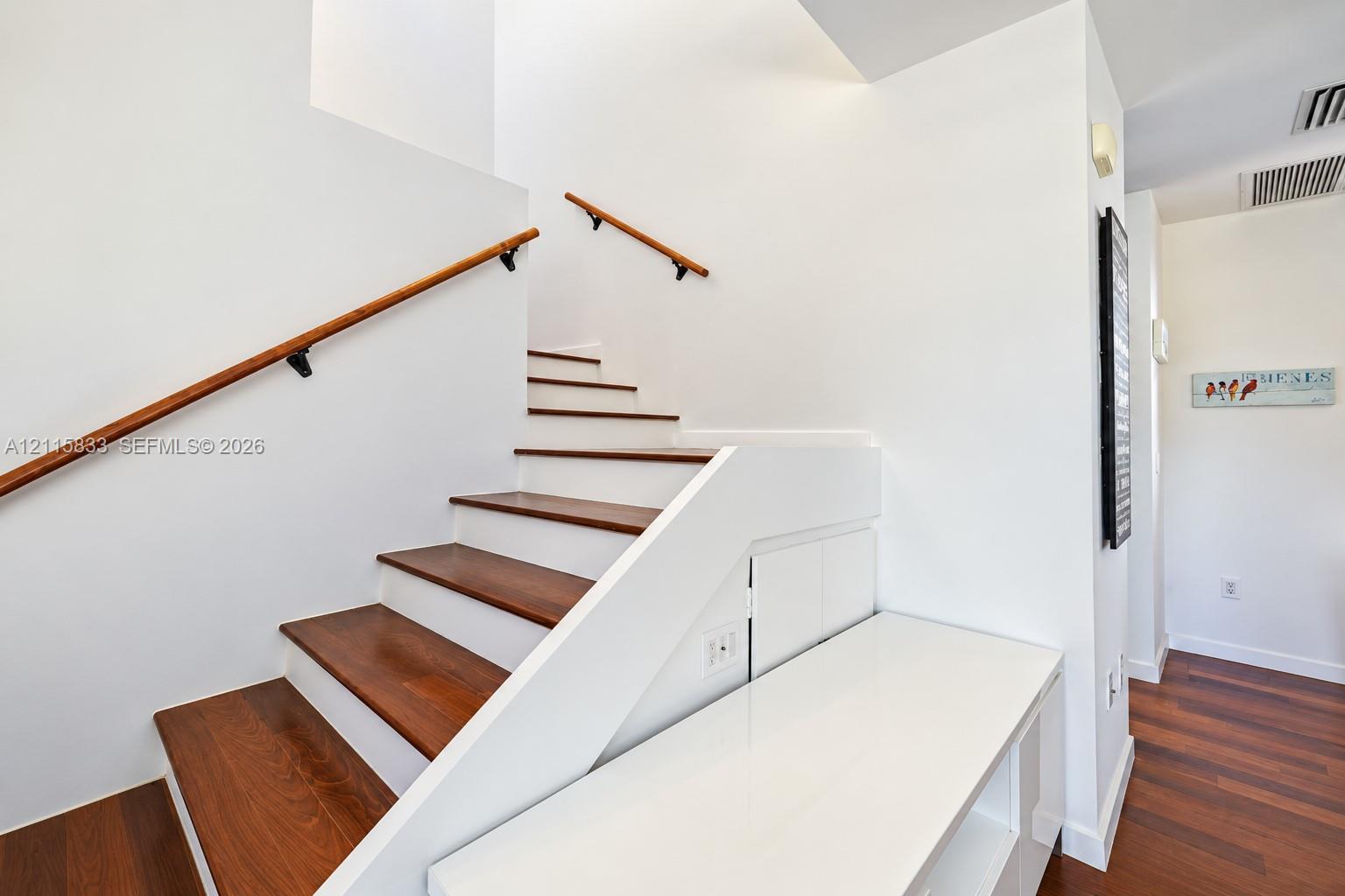 Staircase featuring wood-finish treads, white risers, wooden handrails with black metal brackets, and built-in white under-stair cabinetry