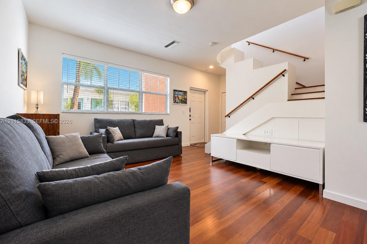 Open-concept interior featuring wood-finish flooring, a white built-in media console, a staircase with a wooden handrail, and large multi-pane windows